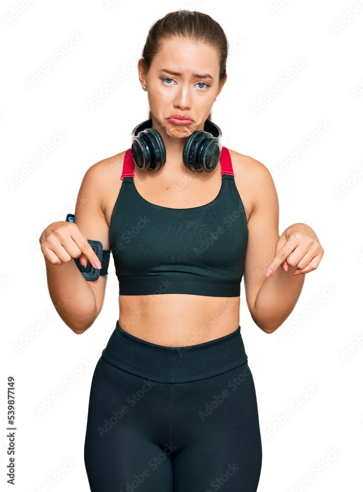 Beautiful blonde woman wearing gym clothes and using headphones pointing down looking sad and upset, indicating direction with fingers, unhappy and depressed. Beautiful blonde woman wearing gym clothes and using headphones pointing down looking sad and upset, indicating direction with fingers, unhappy and depressed.