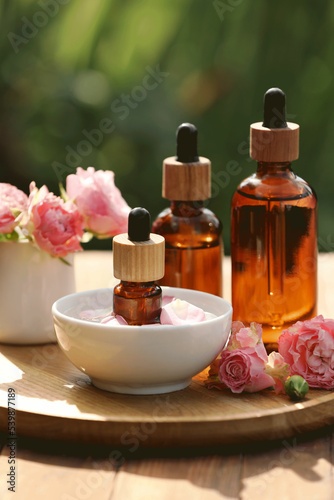 Bottles of rose essential oil and flowers on wooden table outdoors  closeup