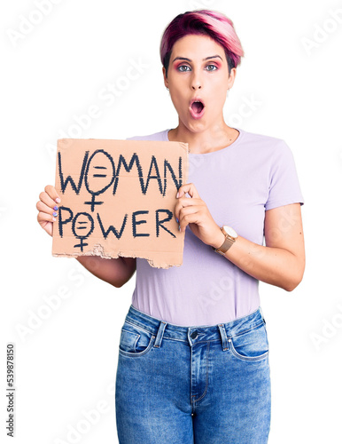 Young beautiful woman with pink hair holding woman power banner scared and amazed with open mouth for surprise, disbelief face © Krakenimages.com