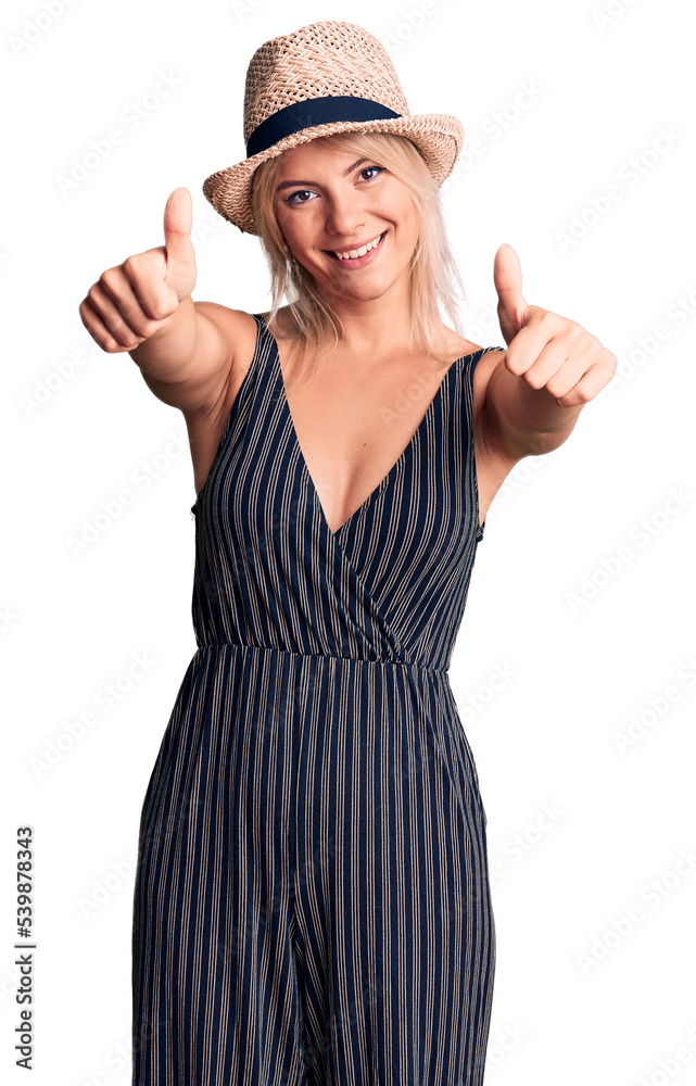 Young beautiful blonde woman wearing summer hat and dress approving doing positive gesture with hand, thumbs up smiling and happy for success. winner gesture. Young beautiful blonde woman wearing summer hat and dress approving doing positive gesture with hand, thumbs up smiling and happy for success. winner gesture.