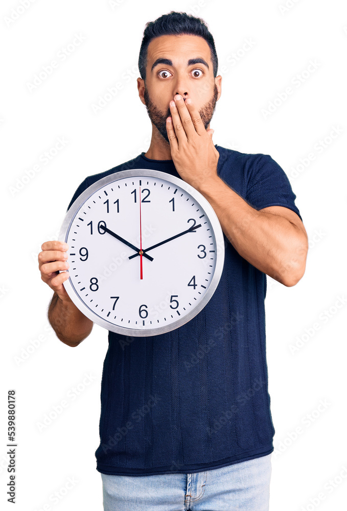 Young hispanic man holding big clock covering mouth with hand, shocked and afraid for mistake. surprised expression Young hispanic man holding big clock covering mouth with hand, shocked and afraid for mistake. surprised expression