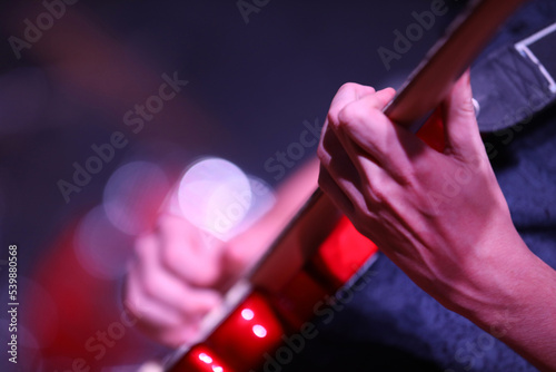 Close up of hand and fingers playing music on a bright red electric guitar. Hand strumming fret with a deliberately blurred spotlight background