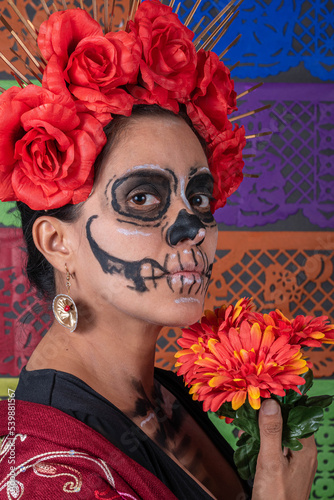Mexican woman in traditional make up and Catrina costume, holding an orange rose. Colorful cut paper background