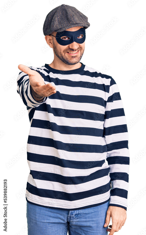 Young handsome man wearing burglar mask smiling cheerful offering palm hand giving assistance and acceptance. Young handsome man wearing burglar mask smiling cheerful offering palm hand giving assistance and acceptance.