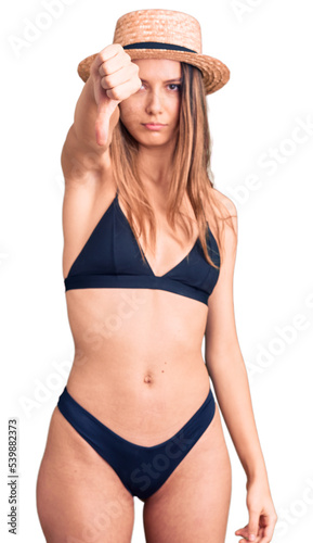 Young beautiful girl wearing bikini and hat looking unhappy and angry showing rejection and negative with thumbs down gesture. bad expression.