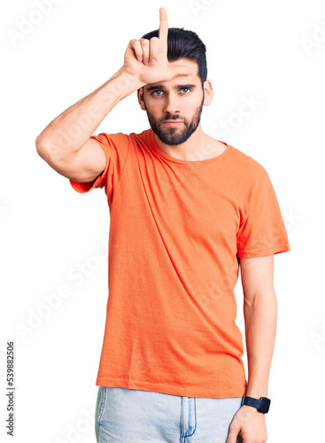 Young handsome man with beard wearing casual t-shirt making fun of people with fingers on forehead doing loser gesture mocking and insulting.