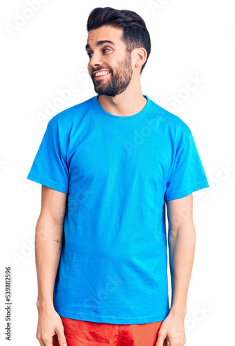 Young handsome man with beard wearing casual t-shirt looking away to side with smile on face, natural expression. laughing confident.