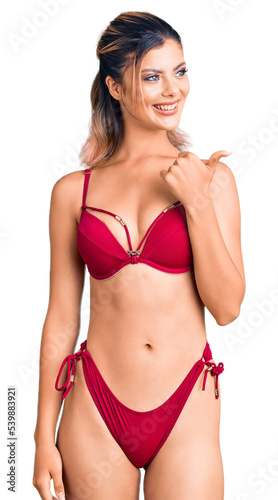 Young beautiful woman wearing bikini smiling with happy face looking and pointing to the side with thumb up. © Krakenimages.com