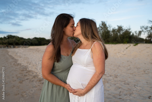 lesbian couple kissing on the beach holding pregnant belly
