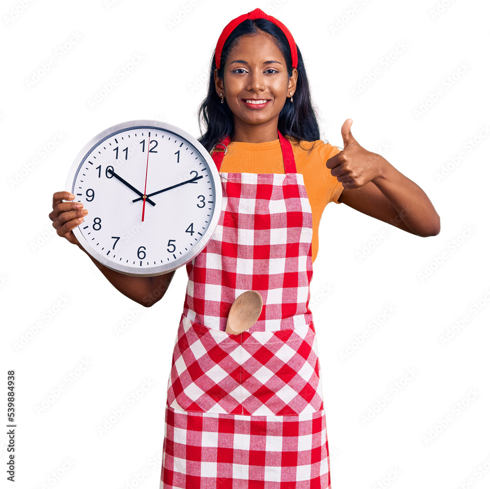 Young indian girl wearing cook apron holding big clock smiling happy and positive, thumb up doing excellent and approval sign Young indian girl wearing cook apron holding big clock smiling happy and positive, thumb up doing excellent and approval sign