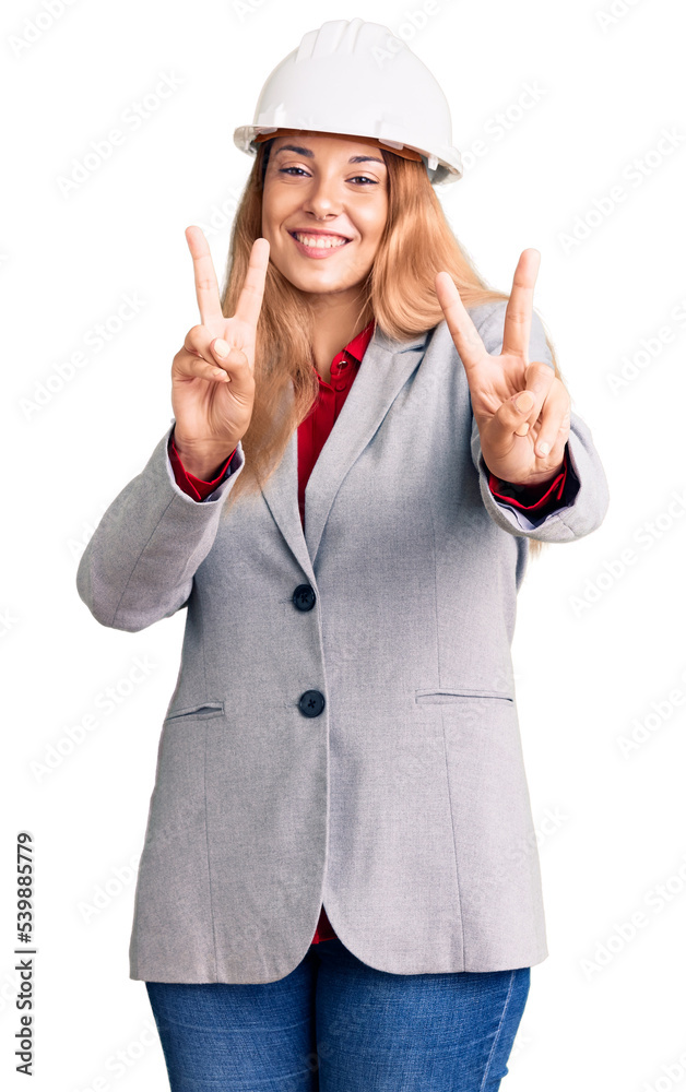 Beautiful young woman wearing architect hardhat smiling looking to the camera showing fingers doing victory sign. number two.