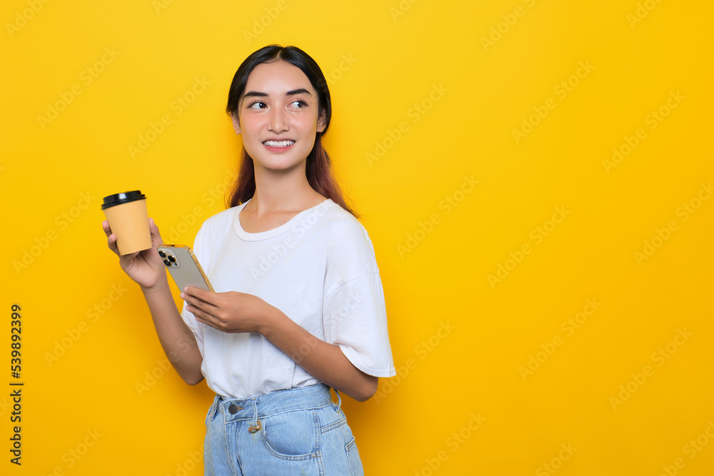 Cheerful pretty young girl in white t-shirt holding mobile phone and coffee cup, looking away at blank space isolated on yellow background Cheerful pretty young girl in white t-shirt holding mobile phone and coffee cup, looking away at blank space isolated on yellow background