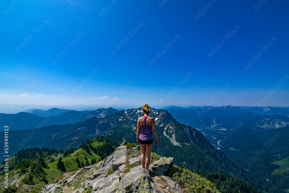 Adventurous athletic woman standing on top of a mountain looking out at a mountain range on a beautiful sunny day in the Pacific Northwest.
