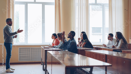 Friendly teacher is talking to students, smart man is raising hand and speaking sitting at table in spacious lecture hall while his classmates are writing.