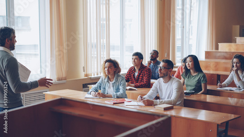 Experienced high school teacher is talking to cheerful students while attractive young people are listening, smiling and laughing sitting at desks in light classroom.