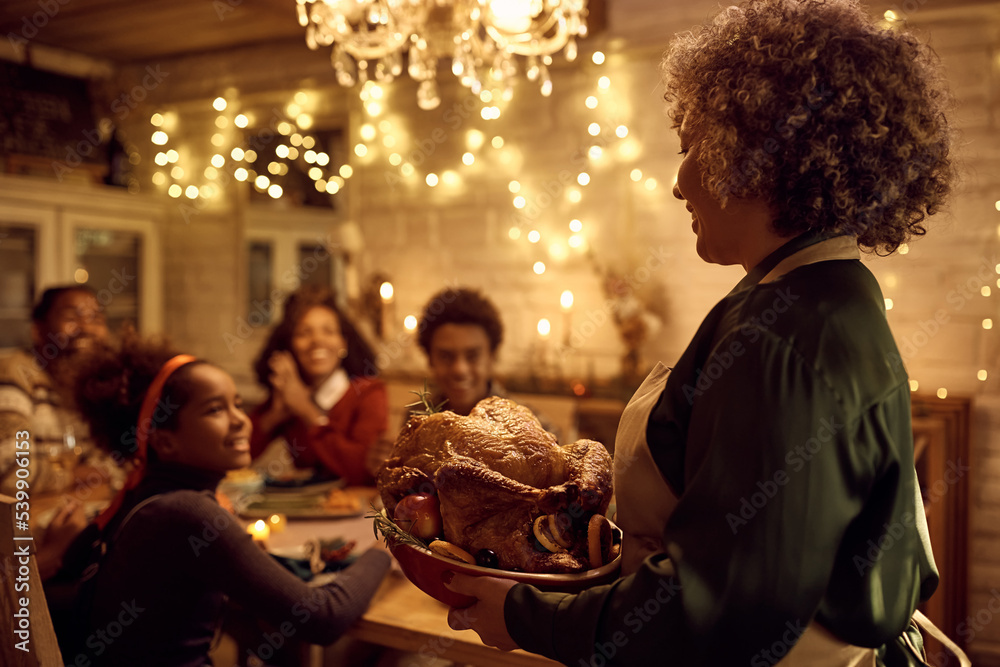 Happy black woman bringing Thanksgiving turkey to dining table during family lunch.