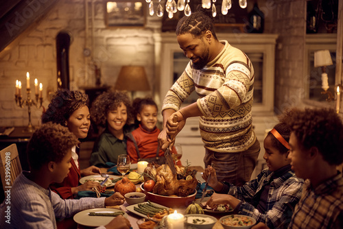 Happy black man carving Thanksgiving turkey during family meal at dining table.