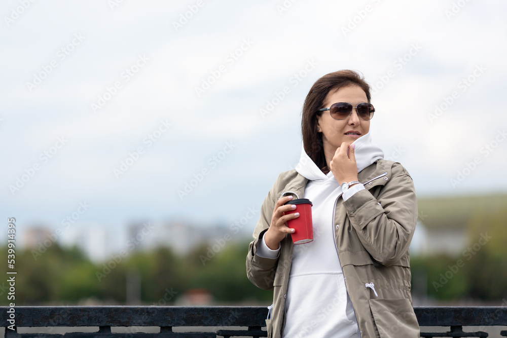 Attractive Caucasian woman wearing dark glasses stands on the embankment outdoor with a coffee in her hand, selective focus. Attractive Caucasian woman wearing dark glasses stands on the embankment outdoor with a coffee in her hand, selective focus.