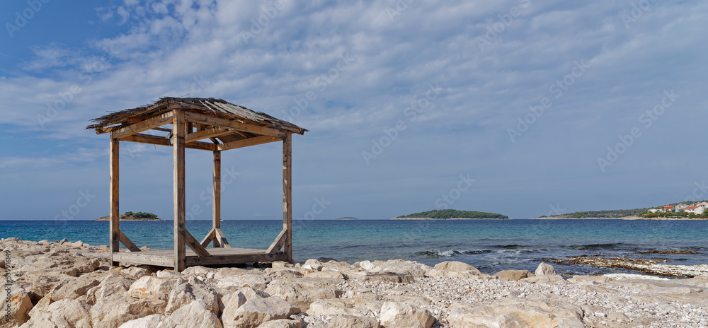Robinson crusoe. Lonely wooden hut covered with reeds on the Croatian Mediterranean coast with blue sea and islands in the background. Copy space for your design or product. Web banner. Robinson crusoe. Lonely wooden hut covered with reeds on the Croatian Mediterranean coast with blue sea and islands in the background. Copy space for your design or product. Web banner.