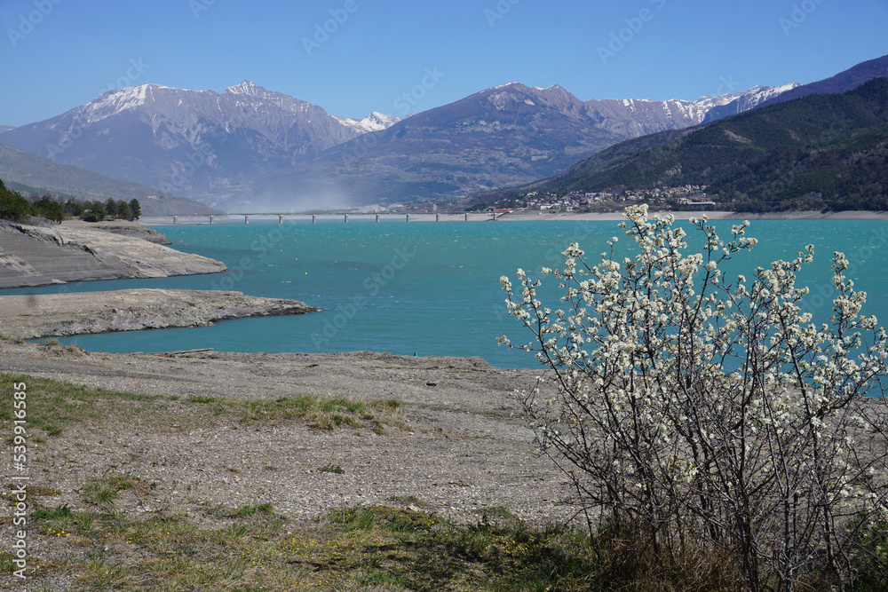panoramic view of the dust storm above embrun from the beach on a spring windy day with blooming white flowers and stone bridge Serre Ponçon lake in the Southern Alps, France 