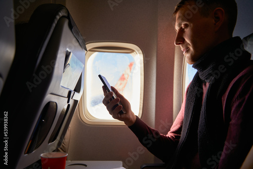 Fotografie Businessman sitting next to airplane window and using smartphone