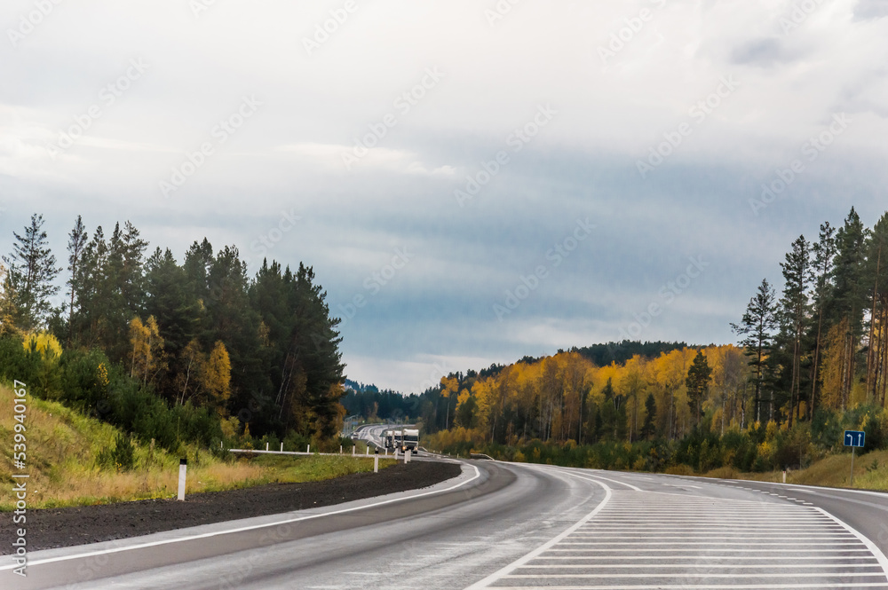 Autumn landscape. On the freeway. Travel by car. White posts on the side of the road and markings with white stripes on the road. Beautiful autumn forest around the road. Nature of Eastern Siberia. Autumn landscape. On the freeway. Travel by car. White posts on the side of the road and markings with white stripes on the road. Beautiful autumn forest around the road. Nature of Eastern Siberia.