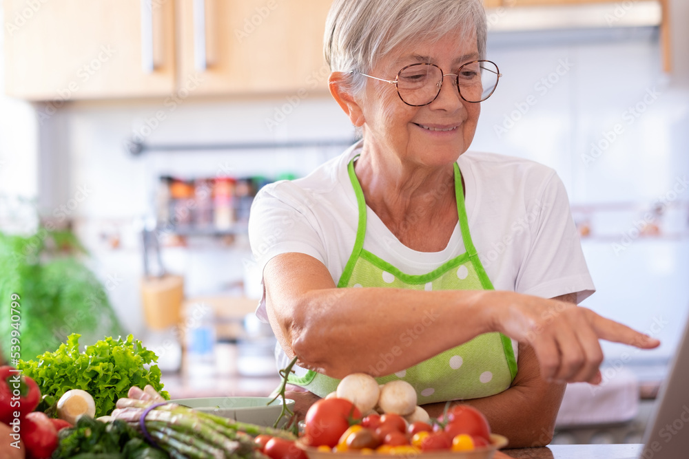 Smiling caucasian senior woman surfing the net sitting in the home kitchen while preparing vegetables. Mature woman using laptop technology looking for new recipes Smiling caucasian senior woman surfing the net sitting in the home kitchen while preparing vegetables. Mature woman using laptop technology looking for new recipes
