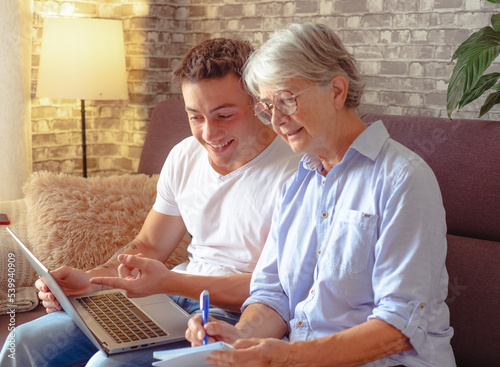 Принти на полотні Smiling young boy sitting on sofa at home helping senior grandmother surf with laptop on the web while she takes notes