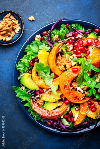 Autumn pumpkin salad with caramelized pumpkin slices, red cabbage, avocado, arugula, pomegranate seeds and walnuts. Healthy vegan, vegetarian eating, comfort food. Blue background. Top view