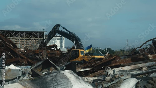 Excavator with Ukrainian flag moving scrap on site of airbombed business center. Aftermath of Russian air strikes. Building destroyed by missile attack
 photo