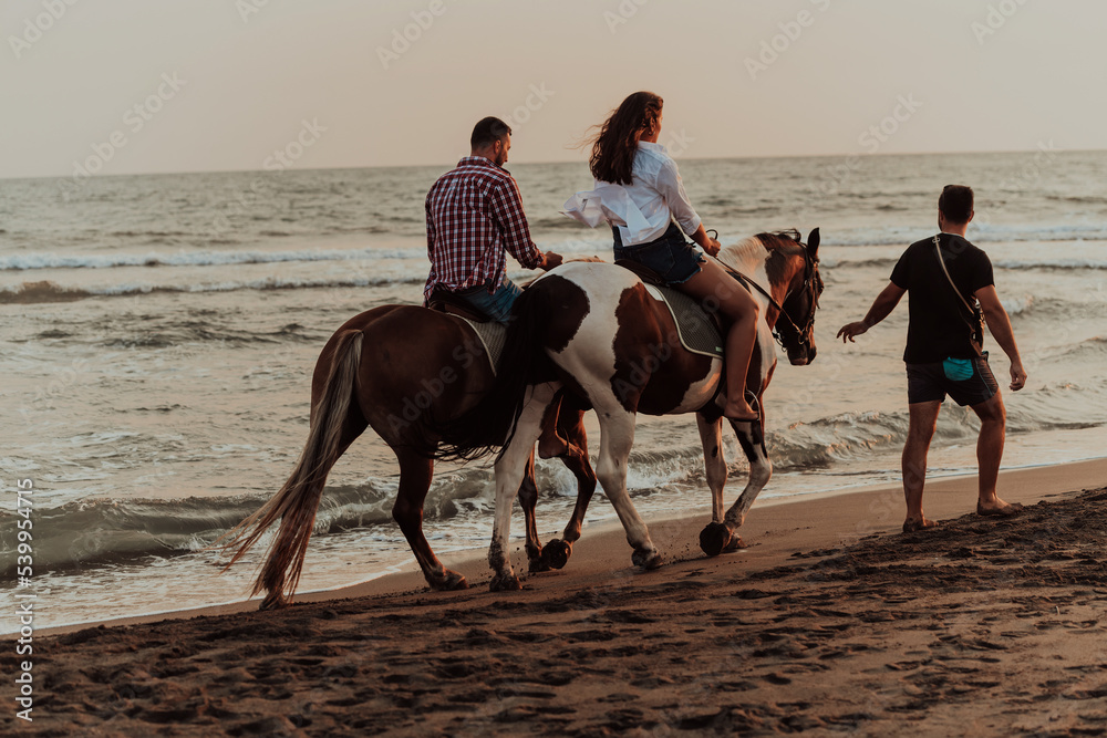 A loving couple in summer clothes riding a horse on a sandy beach at sunset. Sea and sunset in the background. Selective focus 