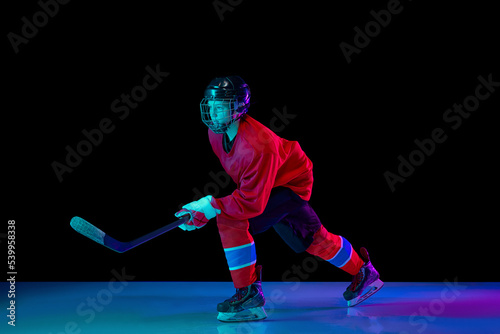 Junior ice hockey player in sports uniform and protective equipment in action over dark background in neon light. Sport, power, challenges, achievement, goals © master1305
