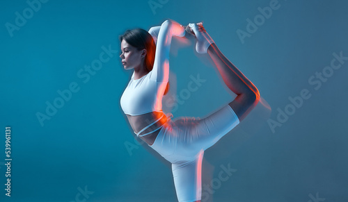 Fototapeta Naklejka Na Ścianę i Meble -  Fitness model doing exercises with motion blur effect. A girl in white sports uniform on blue background stands on one leg and raises the other up.