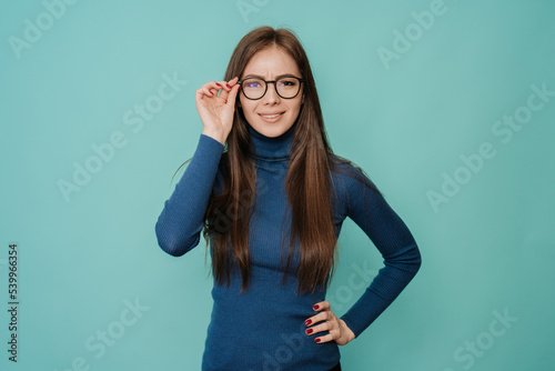 Young beautiful Swedish woman in blue sweater and glasses looking at camera with surprised face expression over turquoise studio background with copy space. Pretty European girl looking with disbelief