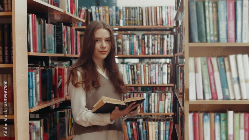 Beautiful student reads a book and then looks at the camera against the background of library Beautiful student reads a book and then looks at the camera against the background of library