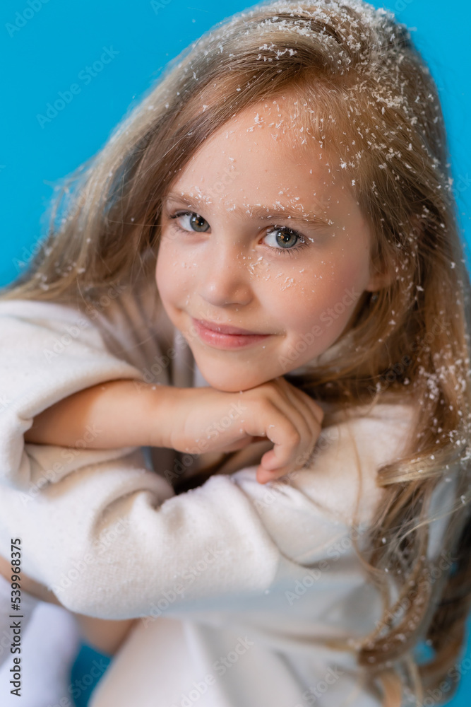 portrait of a cute little blonde girl in a white knitted sweater on a blue background in the studio. snowflakes in the hair, the concept of winter