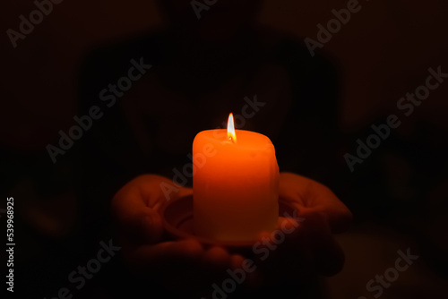 Sad Little boy holding red burning candle in the darkness at home.Ukraine after the war, there is no electricity at home.Selective focus.Closeup.