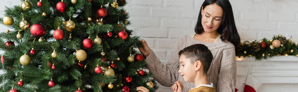 brunette woman smiling near son decorating green spruce with christmas balls, banner brunette woman smiling near son decorating green spruce with christmas balls, banner