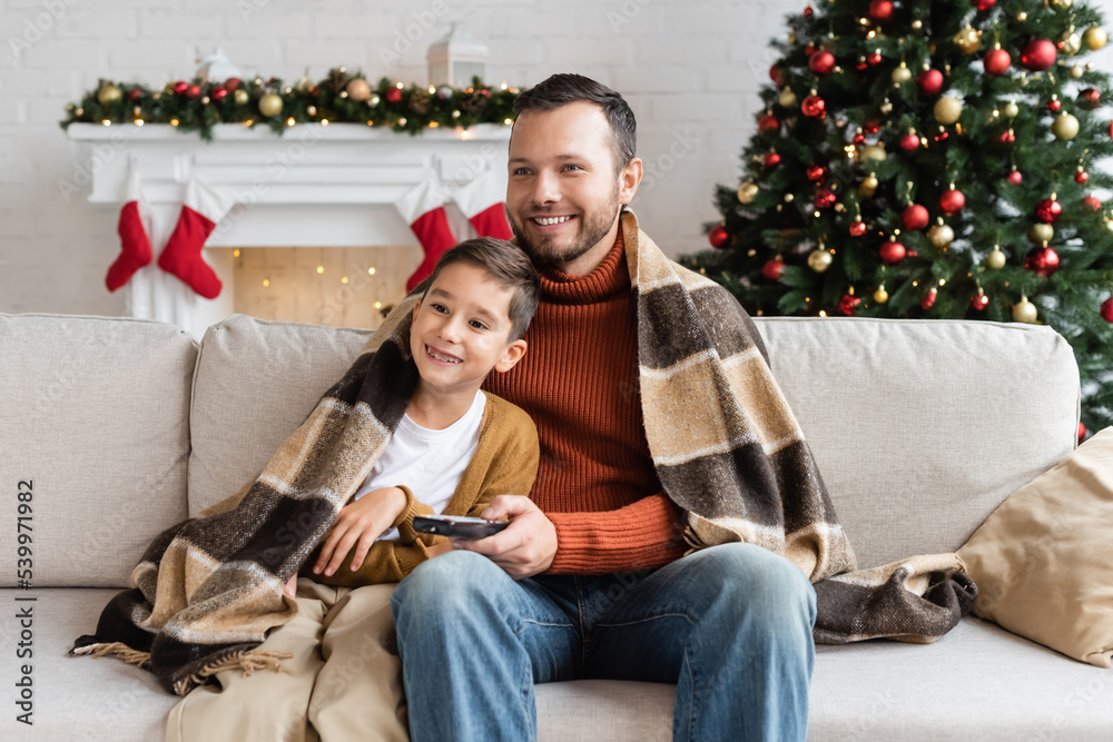 cheerful father and son sitting on couch under warm blanket and watching tv near decorated christmas tree cheerful father and son sitting on couch under warm blanket and watching tv near decorated christmas tree