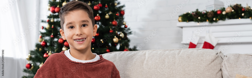 joyful boy in teeth brackets smiling at camera near christmas tree on blurred background, banner