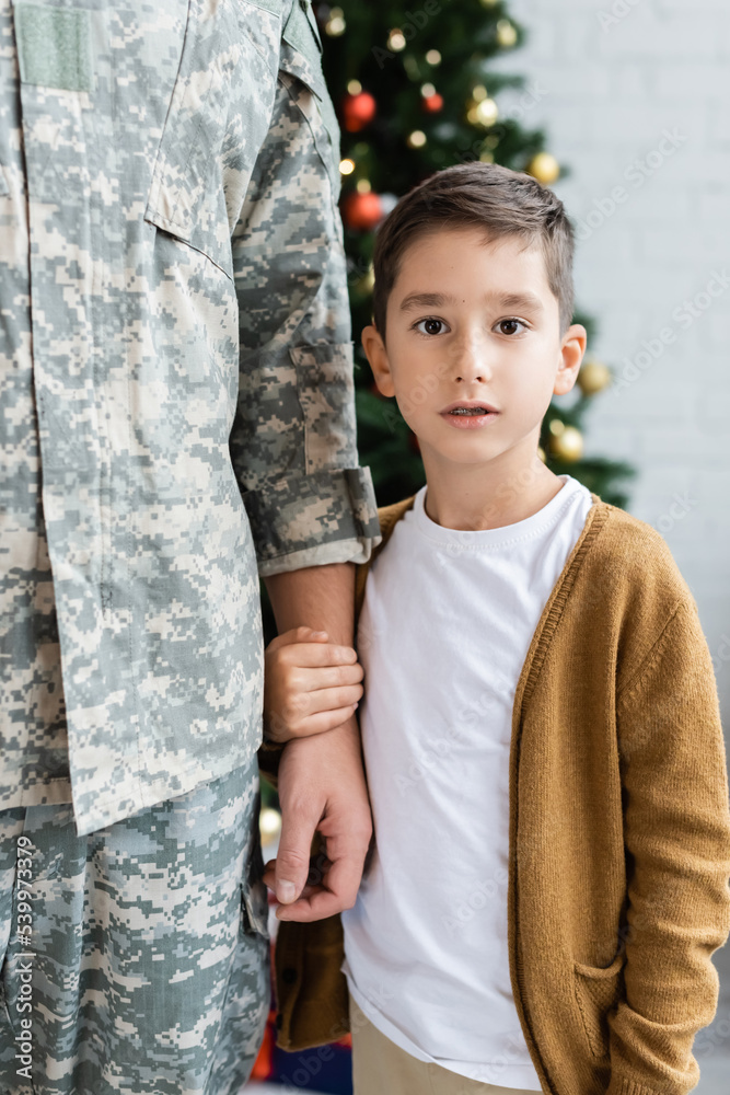 kid looking at camera while holding hand of dad in camouflage at home kid looking at camera while holding hand of dad in camouflage at home