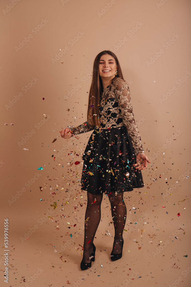 Cheerful smiling girl standing under confetti rain over a beige background. 