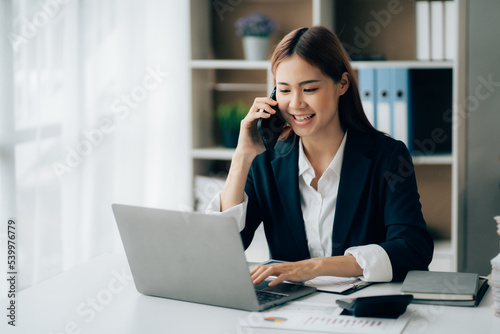 Asian businesswoman talking on phone, using laptop, looking at screen, entrepreneur manager consulting client by call, looking at computer screen, discussing project, reading information