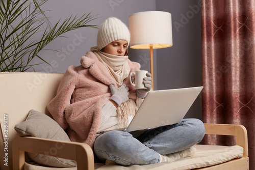 Quadro su tela Profile portrait of woman freelancer in cold room sitting on the sofa wearing warm hat, mittens and cap, works on notebook and trying to get warm while working online on pc computer