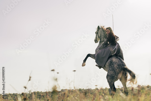 Young woman in image of ancient rider warrior sits with sword on gray rearing up horse on meadow.