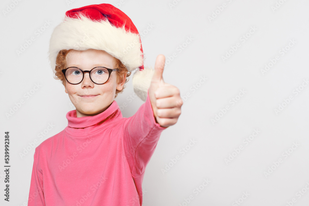 Little redhead boy in Santa hat with thumb up on white background Little redhead boy in Santa hat with thumb up on white background
