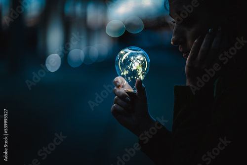 young worried woman holding bright shining lightbulb in her hands feeling unsave while in fear thinking about a future blackout with no electricity or power outage with blue cold background
