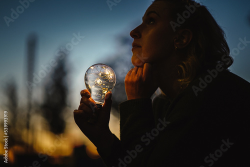 young worried woman holding bright shining lightbulb in her hands feeling unsave while in fear thinking about a future blackout with no electricity or power outage with blue cold background