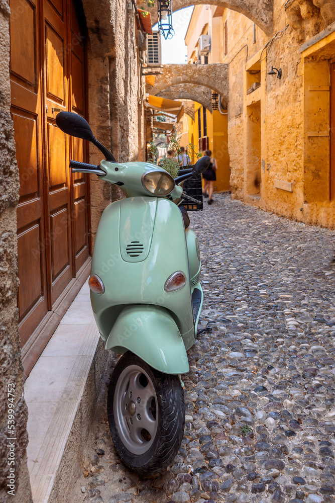 Narrow cobbled historical streets of Rhodes. Tourists walk on Old Town. Vintage scooter bike is parked old stone against wall of house, Rhodes, Dodecanese, Greece