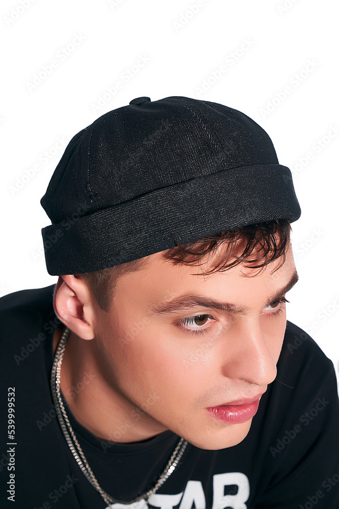 Cropped close-up shot of a young man in a black docker cap with a turn up brim. A guy in a black T-shirt and a denim cap without a visor is posing on a white background. Side view. Cropped close-up shot of a young man in a black docker cap with a turn up brim. A guy in a black T-shirt and a denim cap without a visor is posing on a white background. Side view.